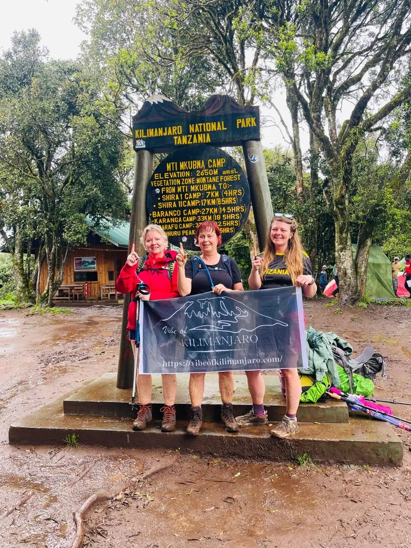 Trekkers holding the Vibe of Kilimanjaro banner at the Mti Mkubwa Camp sign