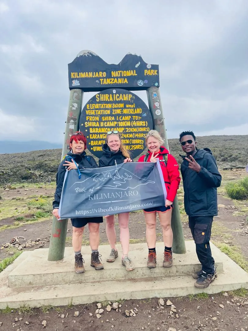 Trekkers and their guide holding the Vibe of Kilimanjaro banner at the Shira 1 Camp sign